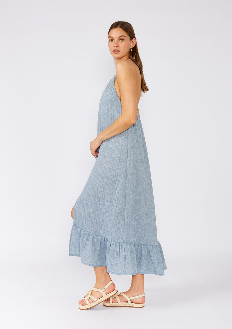 [Color: Dusty Blue] A side facing image of a brunette model wearing a dusty blue bohemian halter beach dress designed in soft cotton gauze. With a v neckline, a long tiered skirt with front slit, an adjustable rope halter tie neckline, and a t back strap detail. 