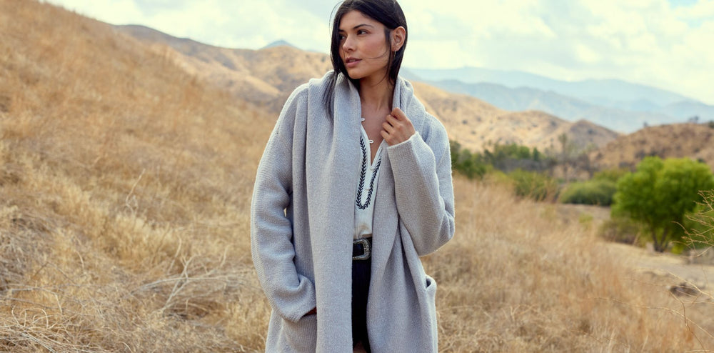 Woman in a long gray coat standing in a dry, open landscape with mountains in the background.
