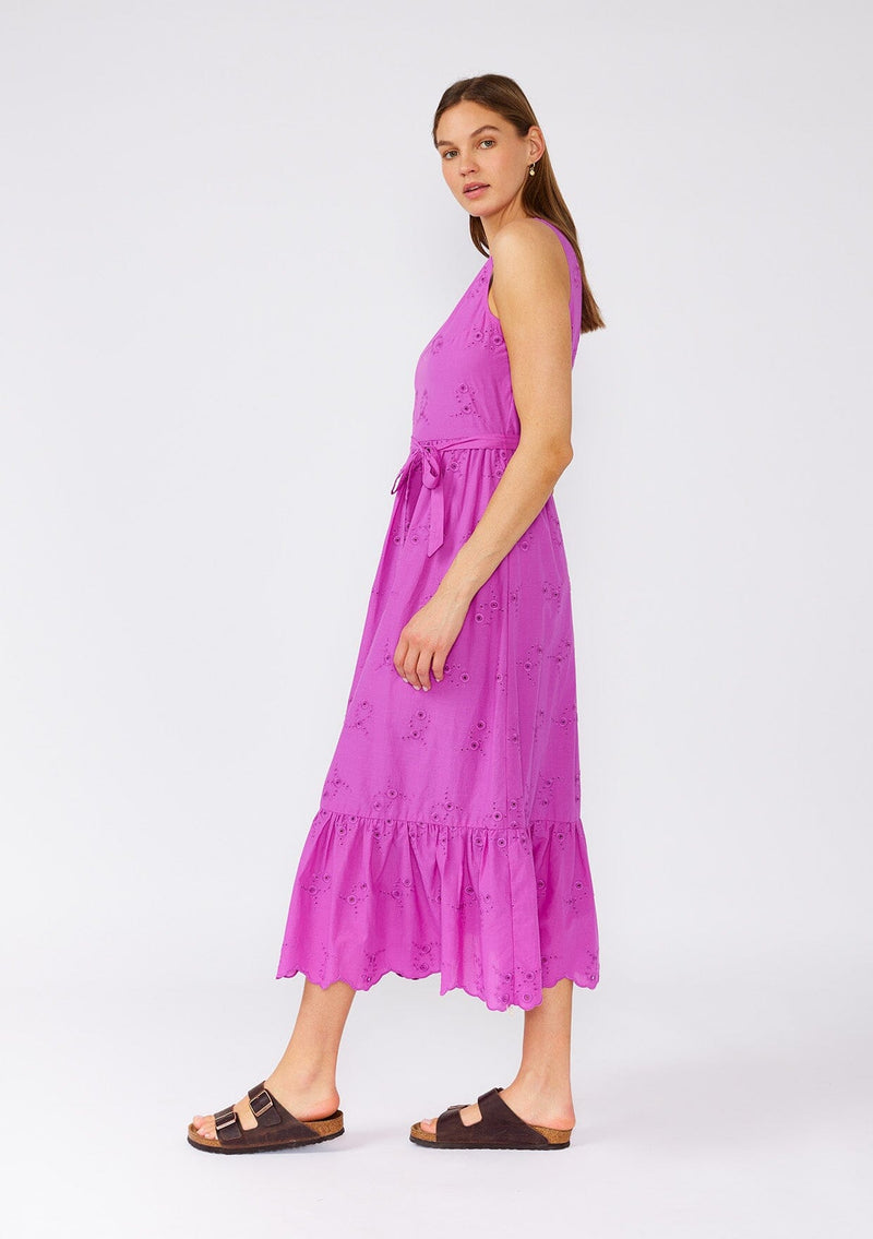 [Color: Orchid] A side facing image of a brunette model wearing a an orchid purple bohemian mid length dress made with embroidered eyelet. A sleeveless spring dress with a v neckline, a tiered skirt, an adjustable waist tie, and a back keyhole with button closure. 