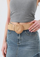 [Color: Tan] A close-up of a model wearing a wide, woven natural tan belt with a large round buckle, paired with a gray ribbed crop top and a blue denim skirt.