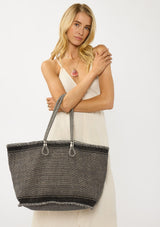 [Color: Black/Natural] A blonde model holding a bohemian tote bag in a natural and black texture. With stitched detail throughout, two rope handles, and a top zip closure.