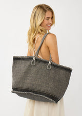 [Color: Black/Natural] A blonde model holding a bohemian tote bag in a natural and black texture. With stitched detail throughout, two rope handles, and a top zip closure.