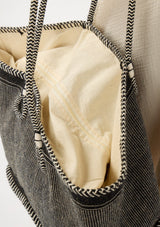 [Color: Black/Natural] A detail image of a bohemian tote bag in a natural and black texture. With stitched detail throughout, two rope handles, and a top zip closure.