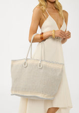 [Color: Blue/Natural] A front facing image of a blonde model holding a bohemian tote bag in a natural and light blue texture. With stitched detail throughout, two rope handles, and a top zip closure.