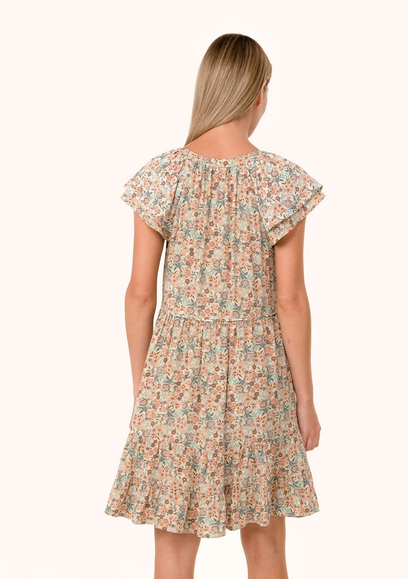 [Color: Cream/Teal] A back facing image of a blonde model wearing a lightweight fall mini dress in a cream and teal floral print. With short double flutter cap sleeves, a tiered skirt, a self covered loop button front, and a v neckline with ties. 
