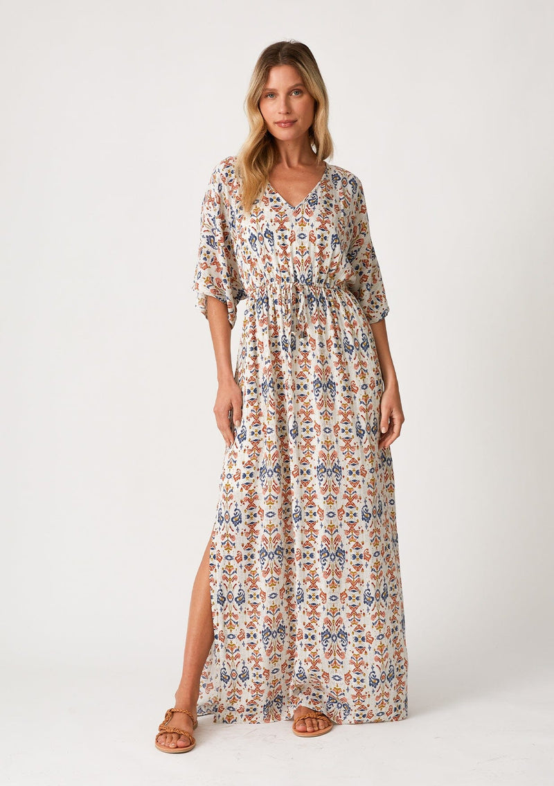 [Color: Off White/Rust] A front facing image of a blonde model wearing a classic maxi dress in a rust red, blue, and off white bohemian print. With short flutter sleeves, a v neckline, a long flowy skirt, side slits, and a drawstring waist with adjustable tie. 