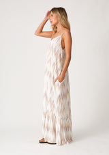 [Color: Natural/Taupe] A side facing image of a blonde model wearing a bohemian maxi tank dress in an off white and taupe chevron striped print. With a v neckline in the front and back, adjustable spaghetti straps, a tiered skirt, side pockets, and a flowy relaxed fit. 
