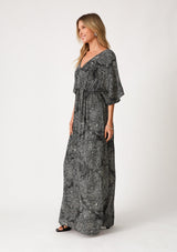 [Color: Black/Natural] A side facing image of a blonde model wearing a flowy bohemian maxi dress in a black floral print. With short sleeves, a v neckline, a long maxi skirt with side slits, and a drawstring waist with adjustable front tie. 