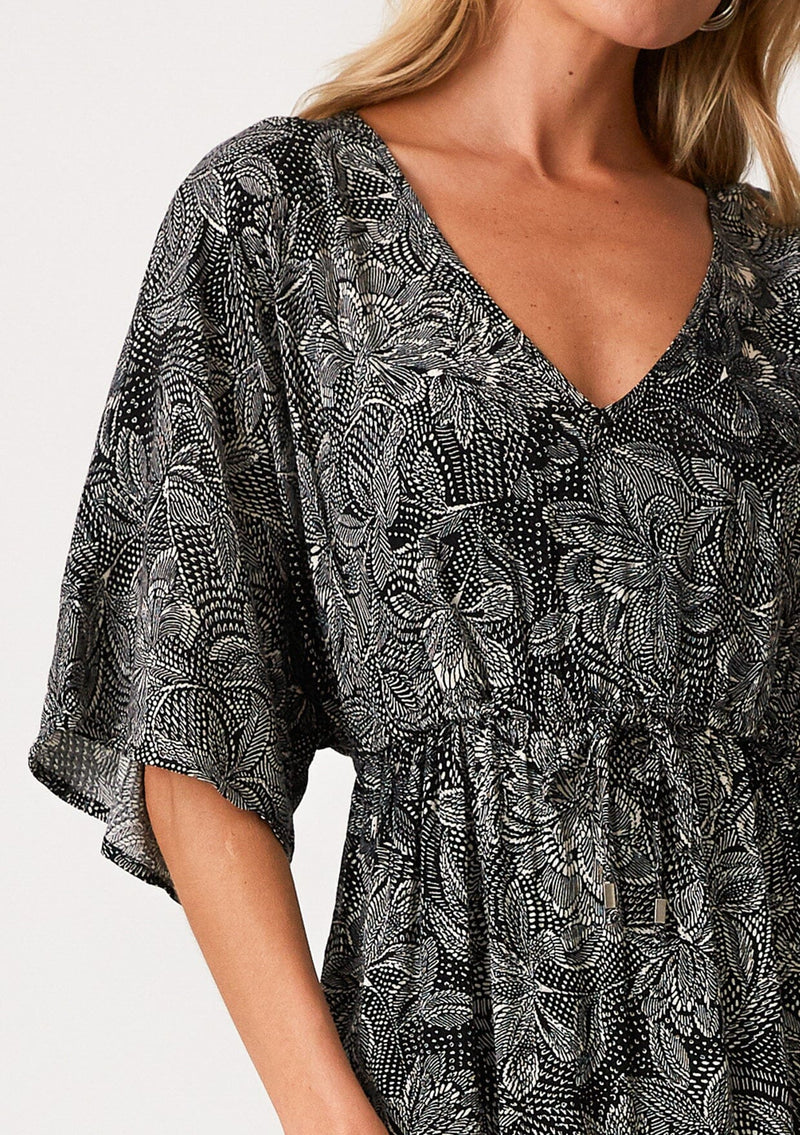 [Color: Black/Natural] A close up front facing image of a blonde model wearing a flowy bohemian maxi dress in a black floral print. With short sleeves, a v neckline, a long maxi skirt with side slits, and a drawstring waist with adjustable front tie. 