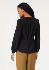 [Color: Black] A back facing image of a brunette model wearing a black blouse with a v neckline, long sleeves, a relaxed fit, and a smocked detail at the back.
