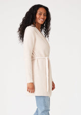 [Color: Petal] A half body side facing image of a brunette model wearing a fuzzy mid length cardigan in soft pink. With long sleeves, a belted waist, and a hoodie. 