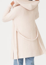 [Color: Petal] A back facing image of a brunette model wearing a fuzzy mid length cardigan in soft pink. With long sleeves, a belted waist, and a hoodie. 