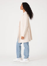 [Color: Petal] A side facing image of a brunette model wearing a fuzzy mid length cardigan in soft pink. With long sleeves, a belted waist, and a hoodie. 