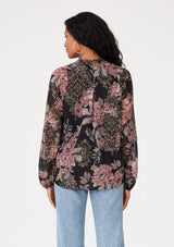 [Color: Black/Dusty Rose] A back facing image of a brunette model wearing a bohemian chiffon blouse designed in a black and pink floral print. With metallic details throughout, a surplice v neckline, long sleeves, and an elastic waist at the front. 
