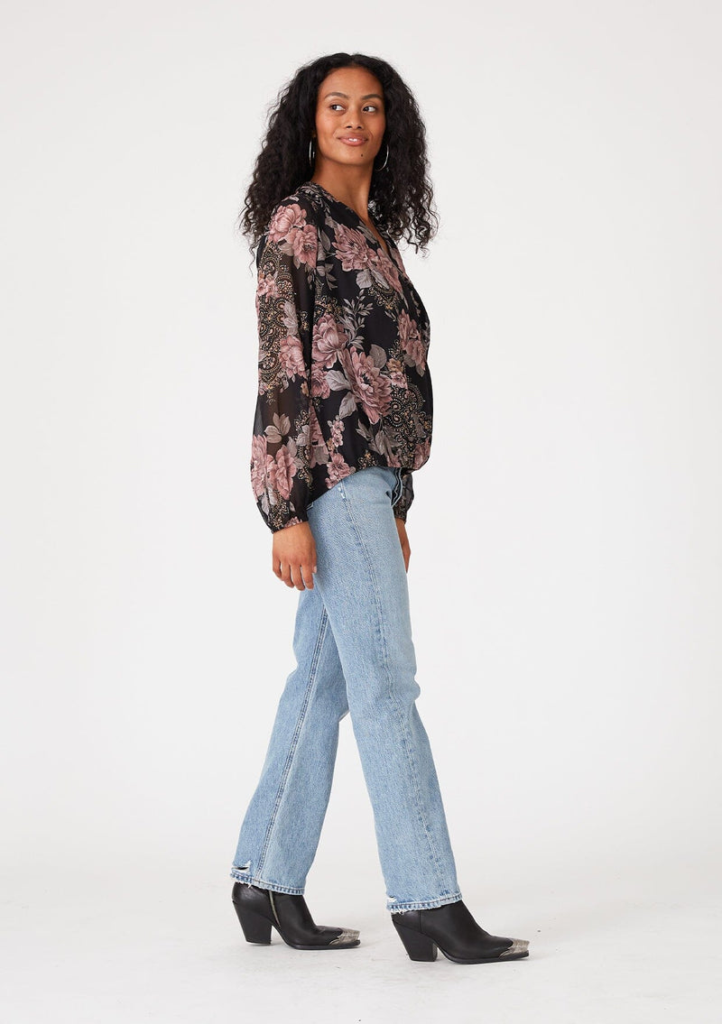 [Color: Black/Dusty Rose] A side facing image of a brunette model wearing a bohemian chiffon blouse designed in a black and pink floral print. With metallic details throughout, a surplice v neckline, long sleeves, and an elastic waist at the front. 