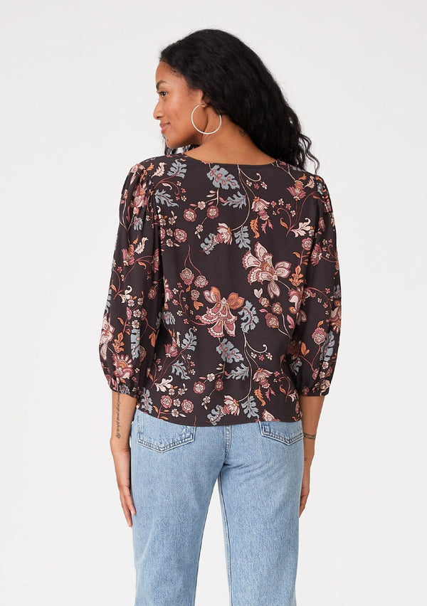 [Color: Brown/Dusty Lilac] A back facing image of a brunette model wearing a bohemian blouse in a brown and dusty purple floral print. With three quarter length sleeves, a v neckline, and a tie front waist. 