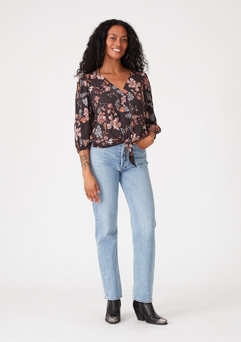 [Color: Brown/Dusty Lilac] A full body front facing image of a brunette model wearing a bohemian blouse in a brown and dusty purple floral print. With three quarter length sleeves, a v neckline, and a tie front waist. 