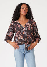 [Color: Brown/Dusty Lilac] A front facing image of a brunette model wearing a bohemian blouse in a brown and dusty purple floral print. With three quarter length sleeves, a v neckline, and a tie front waist. 