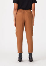 [Color: Rust] A back facing image of a brunette model wearing a rust brown cargo pant with a cropped tapered leg, side pockets, an elastic waist, and a drawstring tie waist.