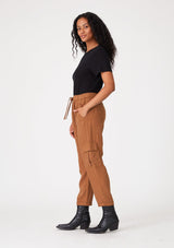 [Color: Rust] A side facing image of a brunette model wearing a rust brown cargo pant with a cropped tapered leg, side pockets, an elastic waist, and a drawstring tie waist.