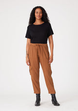 [Color: Rust] A front facing image of a brunette model wearing a rust brown cargo pant with a cropped tapered leg, side pockets, an elastic waist, and a drawstring tie waist. 