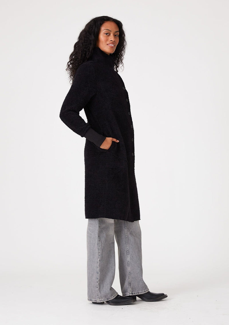 [Color: Black] A side facing image of a brunette model wearing a soft black mid length coat. With long sleeves, a button front, side pockets, and a funnel neckline. 