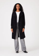 [Color: Black] A full body front facing image of a brunette model wearing a soft black mid length coat. With long sleeves, a button front, side pockets, and a funnel neckline. 