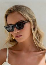 [Color: Tortoise] A blonde woman wearing sleek cat-eye sunglasses with a tortoise frame. 