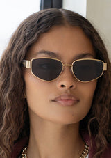 [Color: Gold/Grey] A woman wearing gold metal frame glasses with dark grey lenses from FREYRS. 