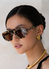 [Color: Milky Tortoise] Woman wearing tortoise print sunglasses and gold jewelry against a white background