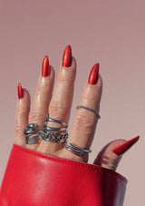 [Color: Scarlet] Hand with red nail polish and silver rings on a pink background