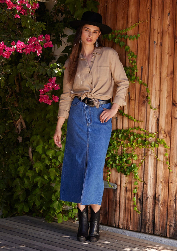 [Color: Sand] A full body front facing image of a brunette model standing outside wearing a relaxed fit khaki shirt. With long sleeves, a collared neckline, a button front, and front patch pockets.