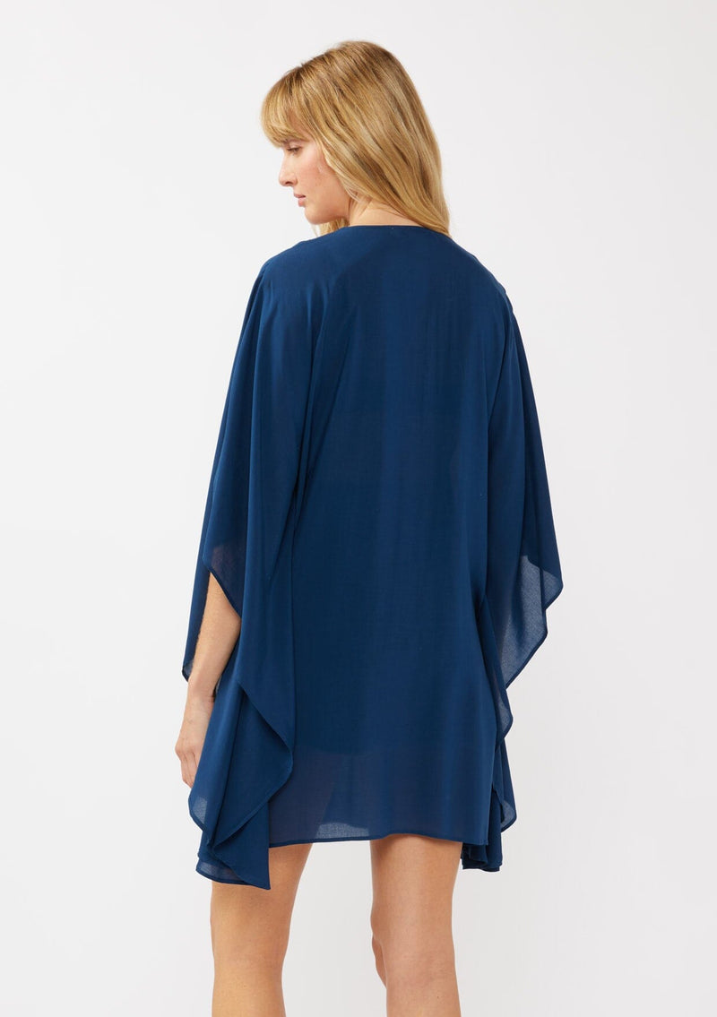 [Color: Navy] A back-facing image of a blonde model wearing a navy embroidered tunic mini dress, perfect as a beach cover-up. This sheer, flowy dress features a split v-neckline with tassel detail and cascading, flowy sleeves.