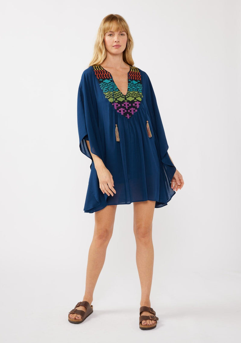 [Color: Navy] A front-facing image of a blonde model wearing a navy embroidered tunic mini dress, perfect as a beach cover-up. This sheer, flowy dress features a split v-neckline with tassel detail and cascading, flowy sleeves.