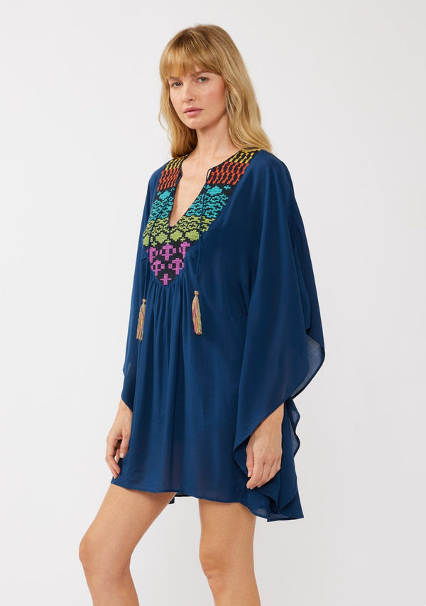 [Color: Navy] A side-facing image of a blonde model wearing a navy embroidered tunic mini dress, perfect as a beach cover-up. This sheer, flowy dress features a split v-neckline with tassel detail and cascading, flowy sleeves.