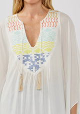 [Color: Off White] A detail image of a blonde model wearing a white embroidered tunic mini dress, perfect as a beach cover-up. This sheer, flowy dress features a split v-neckline with tassel detail and cascading, flowy sleeves.