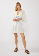 [Color: Off White] A front-facing image of a blonde model wearing a white embroidered tunic mini dress, perfect as a beach cover-up. This sheer, flowy dress features a split v-neckline with tassel detail and cascading, flowy sleeves.