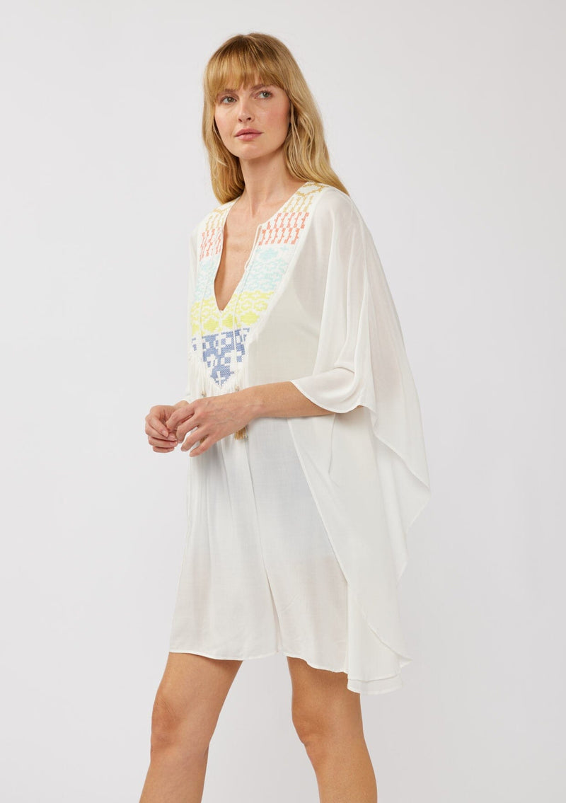 [Color: Off White] A side-facing image of a blonde model wearing a white embroidered tunic mini dress, perfect as a beach cover-up. This sheer, flowy dress features a split v-neckline with tassel detail and cascading, flowy sleeves.