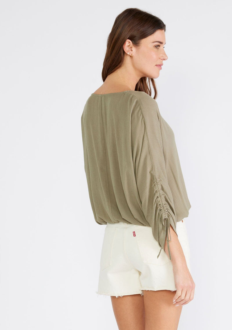 [Color: Olive] A back facing image of a brunette model wearing an olive green bohemian top. With a v neckline, a button front, an elastic waist, and three quarter length sleeves with adjustable ties. 
