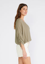 [Color: Olive] A side facing image of a brunette model wearing an olive green bohemian top. With a v neckline, a button front, an elastic waist, and three quarter length sleeves with adjustable ties. 