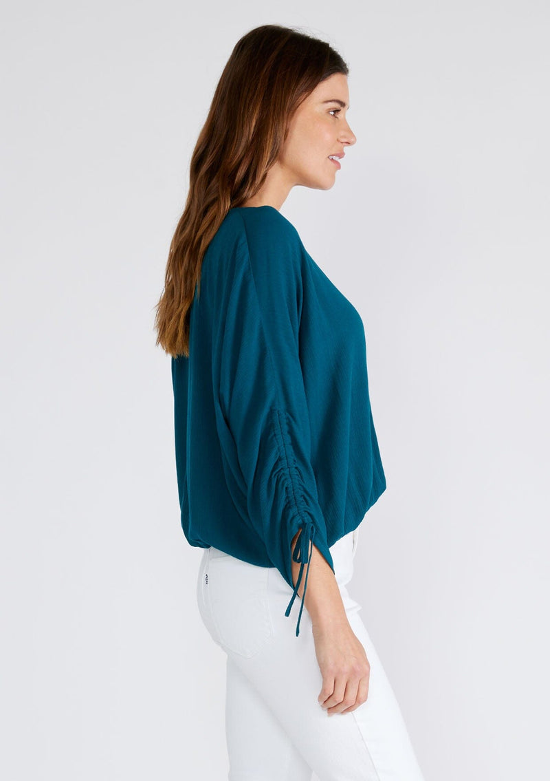 [Color: Peacock] A side facing image of a brunette model wearing a dark teal blue bohemian top. With a v neckline, a button front, an elastic waist, and three quarter length sleeves with adjustable ties. 
