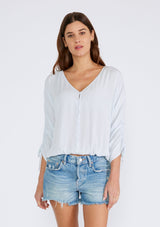 [Color: Ice Blue] A half body front facing image of a brunette model wearing a light blue bohemian top with a button front, a v neckline, three quarter length sleeves with a tie detail, and an elastic waist. 