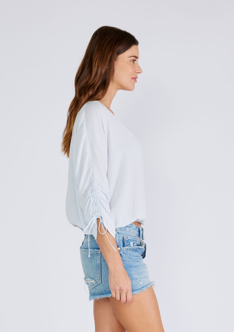 [Color: Ice Blue] A side facing image of a brunette model wearing a light blue bohemian top with a button front, a v neckline, three quarter length sleeves with a tie detail, and an elastic waist. 