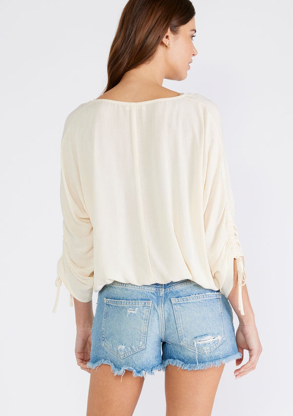 [Color: Ivory] A back facing image of a brunette model wearing an ivory bohemian top with a button front, a v neckline, three quarter length sleeves with a tie detail, and an elastic waist. 