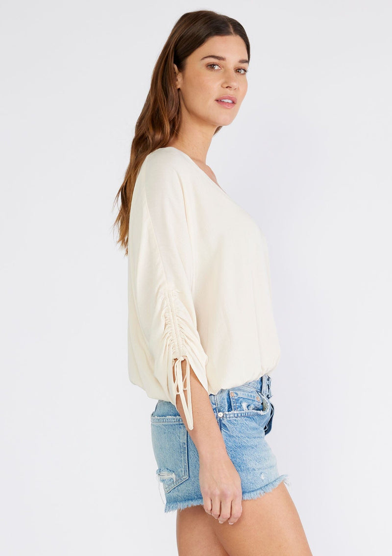 [Color: Ivory] A side facing image of a brunette model wearing an ivory bohemian top with a button front, a v neckline, three quarter length sleeves with a tie detail, and an elastic waist. 