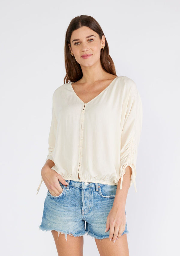 [Color: Ivory] A front facing image of a brunette model wearing an ivory bohemian top with a button front, a v neckline, three quarter length sleeves with a tie detail, and an elastic waist. 