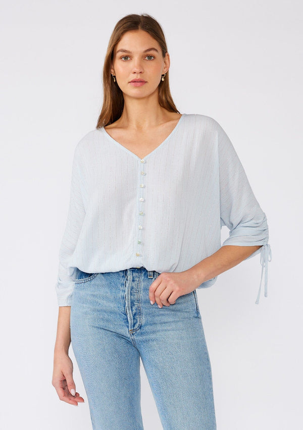 [Color: Periwinkle] A front facing image of a brunette model wearing a light blue bohemian top with gold metallic thread details. With three quarter length sleeves, gathered drawstring sleeve with ties, a v neckline, a button front, and an elastic waist. 