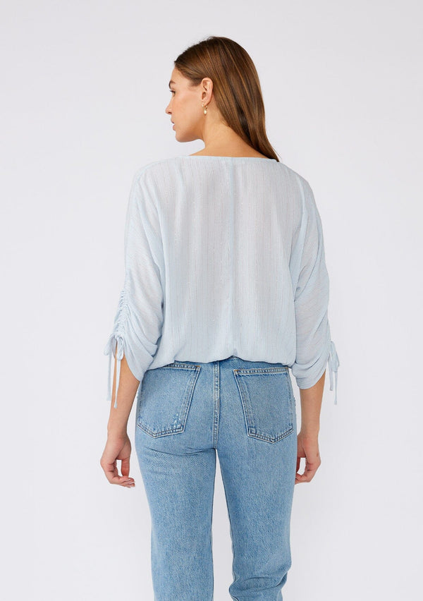[Color: Periwinkle] A back facing image of a brunette model wearing a light blue bohemian top with gold metallic thread details. With three quarter length sleeves, gathered drawstring sleeve with ties, a v neckline, a button front, and an elastic waist. 