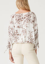 [Color: Ivory/Taupe] A back facing image of a blonde model wearing a lightweight spring top in a white and taupe purple floral print and metallic thread detail. With three quarter length sleeve, a gathered sleeve detail with ties, a v neckline, and a self covered button front. 