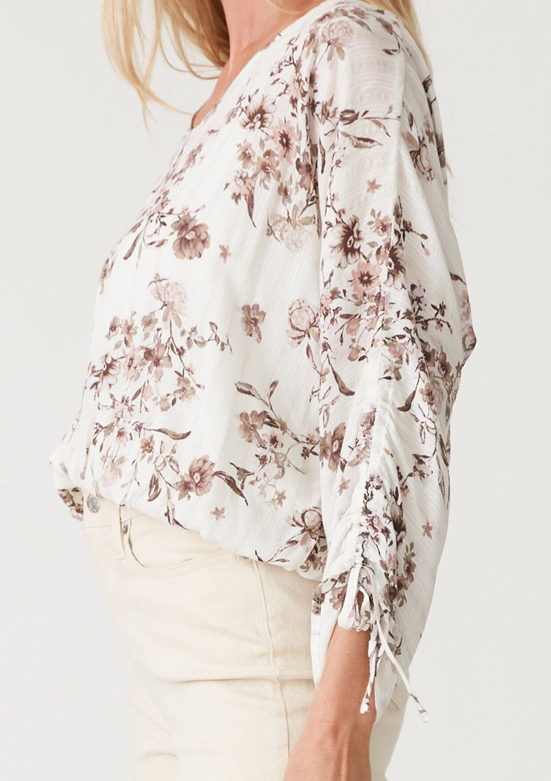 [Color: Ivory/Taupe] A close up side facing image of a blonde model wearing a lightweight spring top in a white and taupe purple floral print and metallic thread detail. With three quarter length sleeve, a gathered sleeve detail with ties, a v neckline, and a self covered button front. 